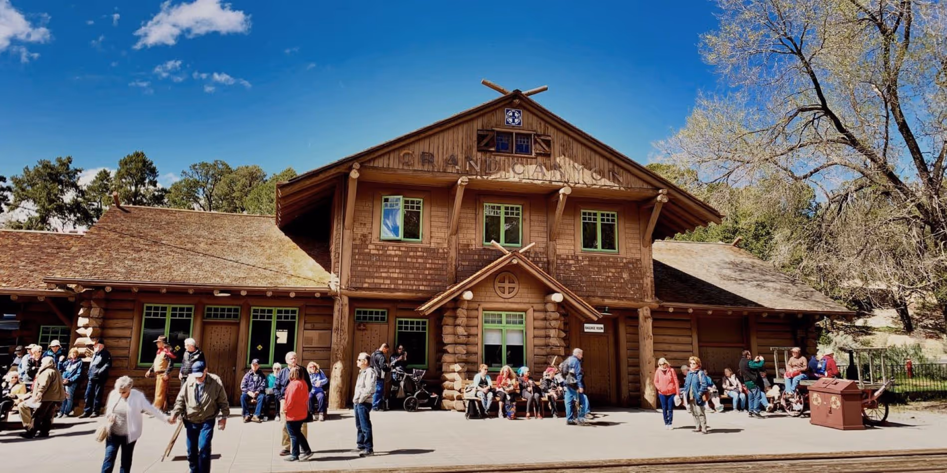 Wooden log cabin building with multiple windows and a peaked roof, surrounded by people sitting and standing under a clear blue sky.
